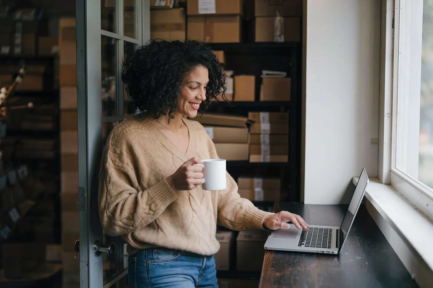 Een webshopeigenaar met een mok koffie aan haar laptop bij het raam van haar magazijn, met stellingen vol kartonnen dozen achter haar